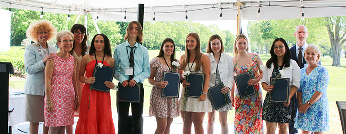 A group of smiling people, including several young adults and an older woman, holding awards or certificates in an outdoor setting with trees in the background.