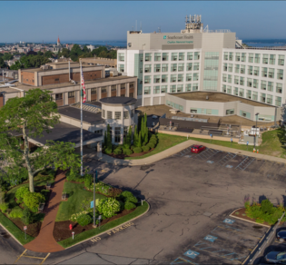Panoramic view of a large beige building surrounded by gardens and parking lots, overlooking a city skyline.
